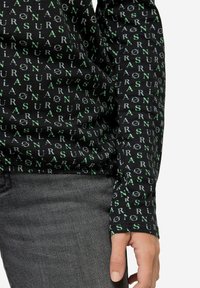Zwarte langemouwen shirt met een herhalend patroon van groene en witte tekst. Zachte stof met een getailleerd ontwerp en mouwen met cuffs.