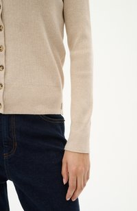 Cardigan beige a coste con chiusura a bottoni, maniche lunghe e un'etichetta sull'orlo, abbinato a jeans in denim blu scuro.