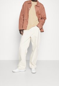 Veste rose légère avec coutures blanches, pull en maille beige, et pantalon ample crème. Complété par des baskets blanches.