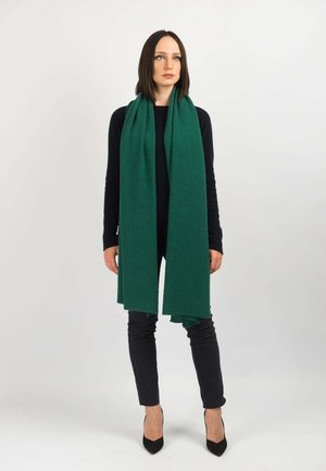 Sciarpa di lana verde con frange, avvolta attorno al collo. Il modello indossa un maglione scuro e pantaloni neri aderenti, posando davanti a uno sfondo bianco.