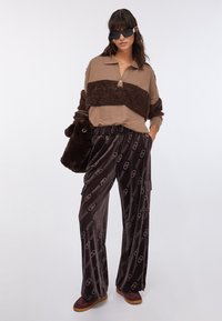 Maglione a righe marroni e beige con trama pelosa, abbinato a pantaloni di velluto scuro con motivo e occhiali da sole rotondi; tiene una piccola borsa di peluche.