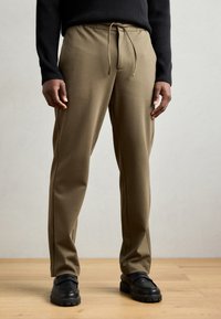 Pantalons khaki texturés avec taille à cordon, coutures latérales et coupe fuselée, associés à des chaussures noires sur un sol en bois.