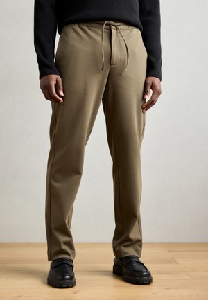 Pantalone u boji khaki, teksturirani, s pojasom na vezanje, bočnim šavovima i suženim krojem, u kombinaciji s crnim cipelama na drvenom podu.