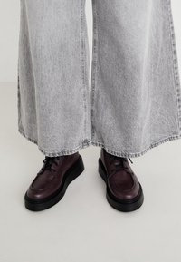 Sapatos de couro burgundy com atacadores e uma sola preta grossa, combinados com calças de denim cinza de perna larga com uma bainha desfiada.
