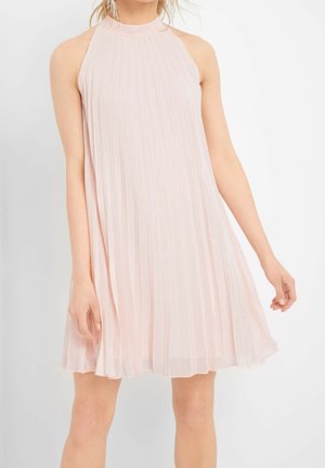 Woman wearing sleeveless, pale pink pleated dress with high neckline and dangling earrings, standing against white background.