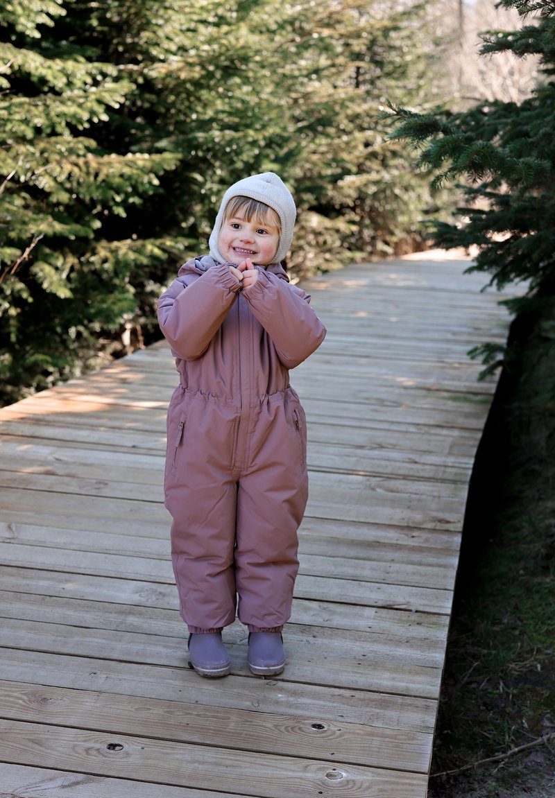 Lapsi vaaleanvioletissa, eristetyssä lumipuvussa, jossa on elastiset hihansuut ja etuvetoketju, harmaa neulottu hattu päässä, seisomassa puisella polulla.