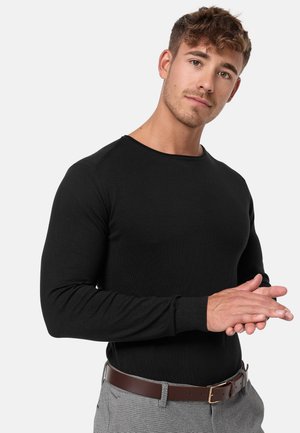 Maglietta nera a maniche lunghe con texture a coste, scollo rotondo e design aderente. Modello che indossa una cintura marrone con pantaloni grigi.