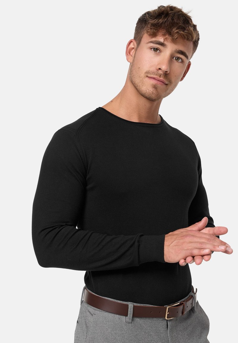 Svart langermet skjorte med ribbet tekstur, rund hals åpning og tettsittende design. Modell iført et brunt belte med grå bukser.