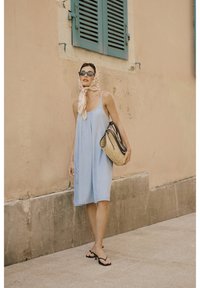 Jasnoniebieska sukienka na ramiączkach, do kolan, z kieszeniami. Pleciona torebka ze słomy i czarne sandały. Jedwabny szalik przewiązany wokół głowy.