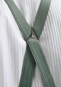 Tirantes verdes con textura y patrón de espiga, con un ajustador de metal plateado, llevados sobre una camisa blanca con plisados.