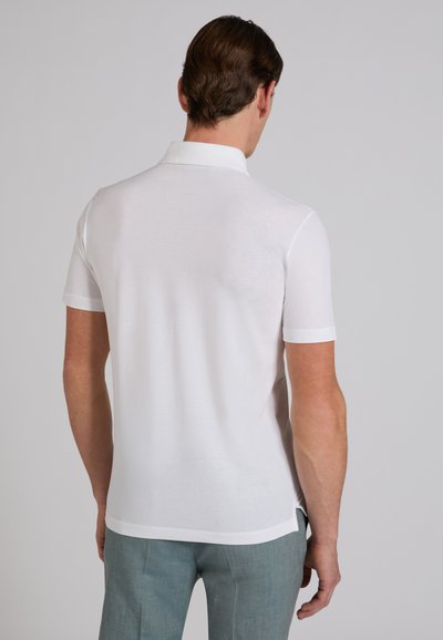 Homme vu de dos portant un polo blanc uni à manches courtes et un pantalon bleu clair sur un fond gris uni.