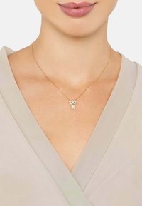 Collar de oro con un colgante de oso de peluche, con una superficie texturada y pequeños adornos, llevado sobre una blusa de color claro.