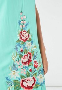Vestido verde menta con intrincados bordados florales en rojo, rosa y blanco. El diseño incluye rosas, hojas y otras plantas.