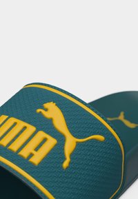 Τιρκουάζ σανδάλι slide με υφασμένο λουρί που διαθέτει ανυψωμένο κίτρινο λογότυπο Puma και όνομα μάρκας με έντονα γράμματα.