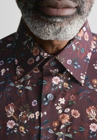 Bloemmotief shirt in donkerbordeaux stof, met verschillende bloemontwerpen in roze, wit en groen, voorzien van grijze knopen en een klassieke kraag.