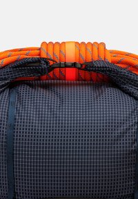 Rygsekk har et navy rutemønster, oransje teksturerte håndtak og en svart clasp. Designet for holdbarhet med forsterkede sømmer.