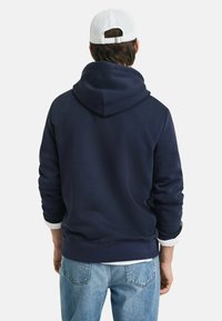 Marineblaue Kapuzenpulli mit weicher Textur, ausgestattet mit einer großen Kapuze und gerippten Bündchen. Kombiniert mit hellblauen Jeans für lässige Kleidung.
