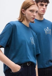 Μπλε βαμβακερά μπλουζάκια με κοντά μανίκια και ριμπ λαιμόκοψη. Στο μπροστινό μέρος υπάρχει το λογότυπο "CALVIN KLEIN" σε λευκό. Φοριέται με σκούρο τζιν παντελόνι.