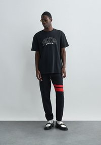 Schwarzes GCDS-Logo-Shirt, lässige Passform, kombiniert mit schwarzen Sweatpants mit roten Streifen und schwarz-weißen Schuhen mit metallischen Akzenten.