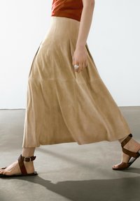 Gonna maxi in suede beige con pieghe, caratterizzata da una texture liscia. Abbinata a sandali in pelle marrone con dettaglio a fibbia.