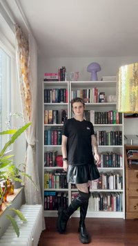 T-shirt nero a maniche corte con taglio dritto, abbinato a una mini gonna in pelle nera lucida e stivali neri alti fino alla coscia, in una stanza piena di libri.