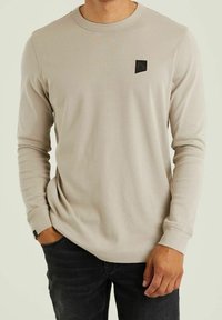 Beige long-sleeve shirt with a ribbed texture, round neckline, and a small black logo patch on the chest, paired with dark jeans.