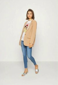 Blazer bege com um botão, usado sobre uma t-shirt gráfica branca, combinado com jeans skinny azuis e sandálias prateadas.