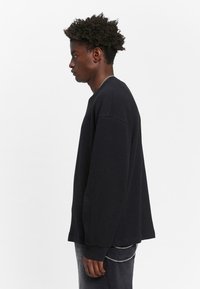Svart langermet sweatshirt i vaffelmønster med løs passform, rund hals og ribbestrikkede mansjetter. Har en sidesplitt og teksturert stoff.