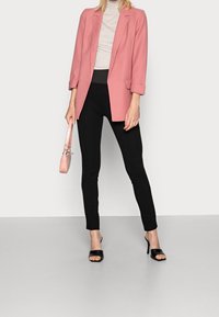 Blazer de suave tejido rosa, corte relajado, con abertura frontal. Suéter de cuello alto gris, leggings negros ajustados y tacones abiertos negros. Bolso rosa con asa.