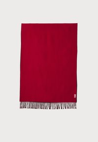 Cobertor de lã vermelho com franjas pretas e uma pequena etiqueta de marca no canto inferior. Textura suave, forma retangular.
