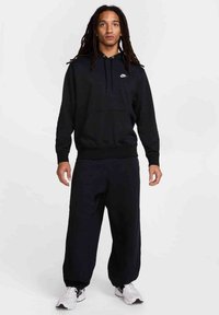 Felpa nera in cotone con tasca frontale e cordino, abbinata a pantaloni della tuta neri larghi e scarpe da ginnastica bianche.