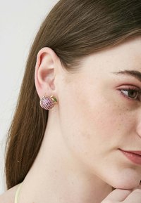 Orecchino a forma di mela, ricoperto di rhinestone rosa con dettagli in oro e una foglia verde, indossato da una persona su uno sfondo neutro.