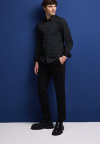 Černá košile s potiskem a úzkým střihem, spárovaná s černými kalhotami a robustními černými botami. Pozadí je modré.