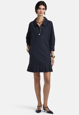 Žena oblečená v tmavomodrých šatách s golierom a skladaným lemom, s trojštvrťovými rukávmi, čiernymi sandálmi s remienkom a zlatými šperkami, stojaca na bielom pozadí.