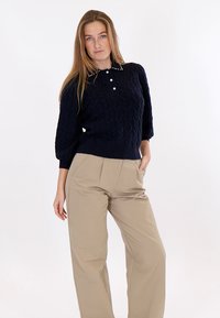 Donna che indossa un maglione blu navy a trecce con bottoni di perle e borchie sul colletto, abbinato a pantaloni beige a gamba larga, in piedi con la mano in tasca.