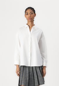 Hvit skjorte med krage, lange ermer og rett snitt. Kombinert med en plissert grå skjørt. Glatt tekstur, minimal design.