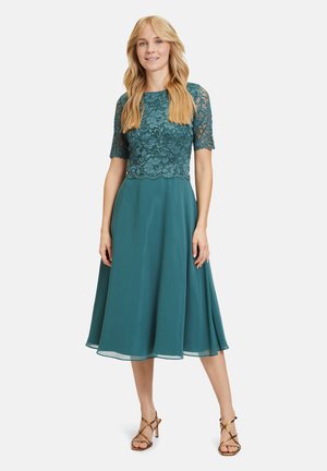 Robe en dentelle bleu sarcelle avec des manches courtes, un corsage ajusté et une jupe évasée. Le tissu est lisse, avec un détail en dentelle festonnée au niveau de la taille.