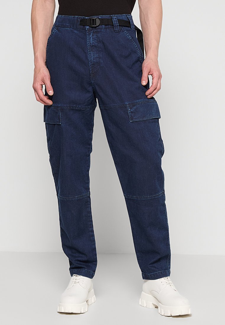 Man in een donkerblauwe cargobroek met een riem en witte chunky schoenen, staand tegen een effen lichte achtergrond.