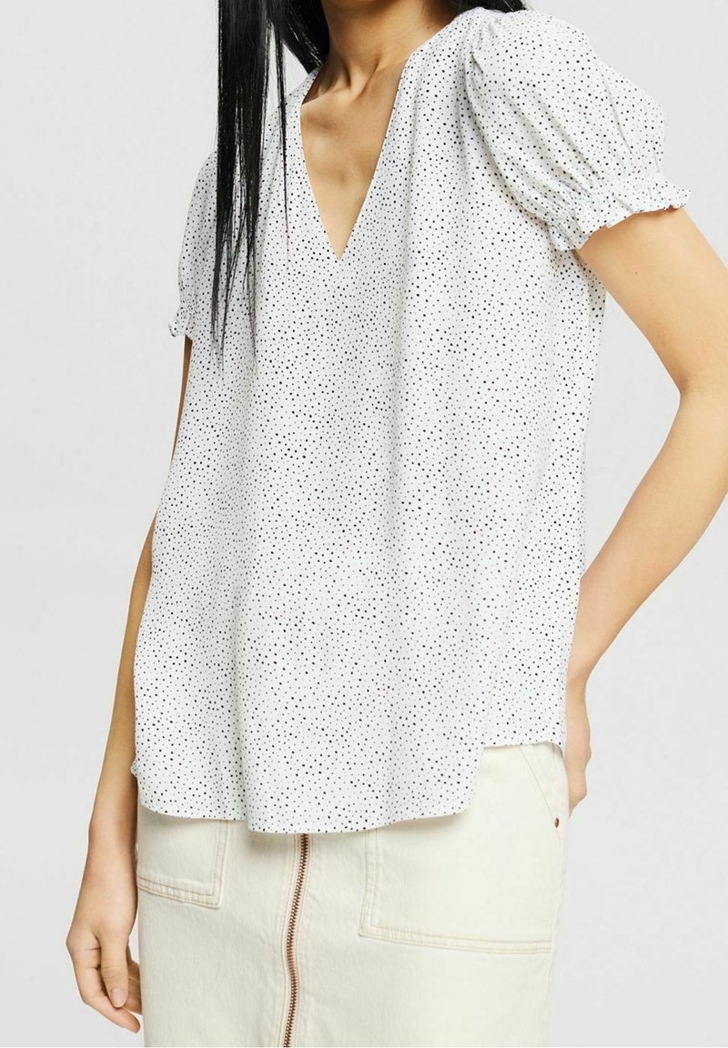 Blusa blanca de manga corta con escote en V, con un patrón de lunares negros. La tela es ligera y presenta detalles fruncidos en las mangas.