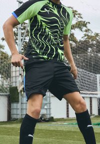 Svart och grön fotbollströja med bladmönster, med korta gröna ärmar och en blå detalj. Svarta shorts och strumpor kompletterar outfiten.