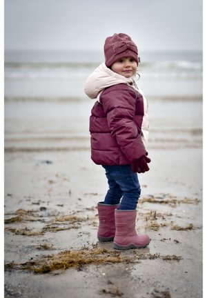 Dziecko ubrane w bordowo-kremową puchową kurtkę, różowe rękawiczki i różowe gumowe kalosze, stojące na piaszczystej plaży blisko wody.