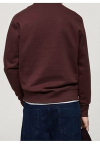 Persoon gekleed in een donkerrode trui met lange mouwen en donkerblauwe spijkerbroek, gezien van achteren, die in één hand een donker object vasthoudt.