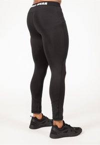 Zwarte sportleggings met een getailleerd ontwerp, een gladde textuur en een contrasterende tailleband met het "Gains Wear" logo. Draagt zwart sportschoenen.