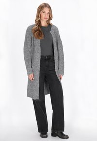 Helle graue Strickjacke aus weichem, strukturiertem Material; langes, offenes Design mit gerippten Kanten. Kombiniert mit dunkel schwarzen, hochgeschnittenen Hosen.