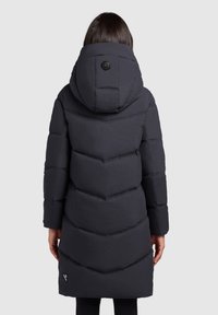 Zwarte pufferjas met een chevron-quiltpatroon, een grote capuchon en een enkele knoopdetail bij de hals. Lange mouwen en een lengte tot halverwege het bovenbeen.