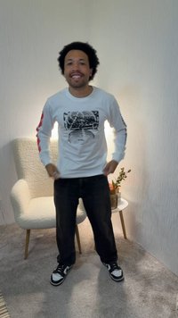Άνδρας με σγουρά μαλλιά που χαμογελάει, φοράει ένα λευκό γραφικό πουκάμισο με μακριά μανίκια και μαύρο παντελόνι, στέκεται σε δωμάτιο με μοκέτα με καρέκλα και έπιπλο πλευράς.