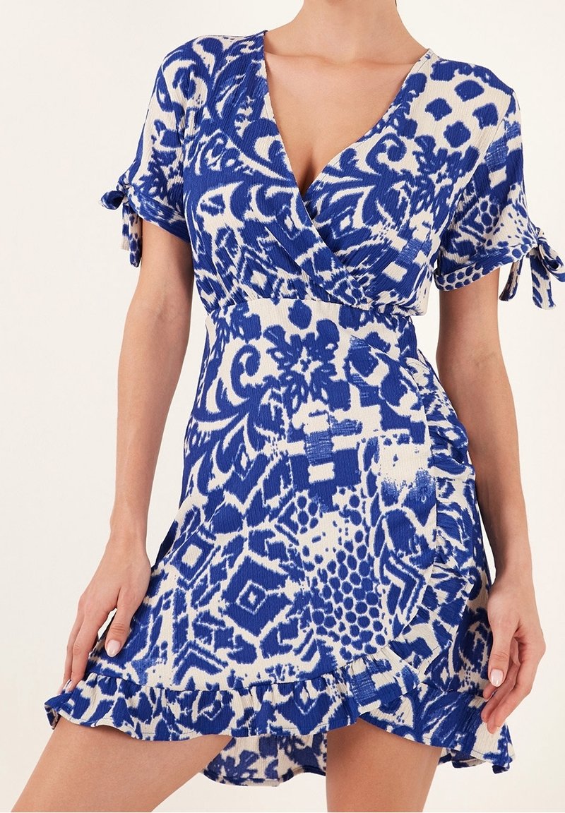 Robe portefeuille courte à motifs bleus et blancs, manches courtes nouées aux poignets et ourlet à volants, portée par une personne.