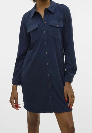 Femme portant une robe-chemise bleu marine à manches longues avec deux poches poitrine, se tenant devant un fond blanc uni.