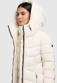 Nafukovacia biela bunda s veľkou, štruktúrovanou kapucňou. Obsahuje zips a kontrastné čierne akcenty, ktoré zdôrazňujú prešívanú textúru.
