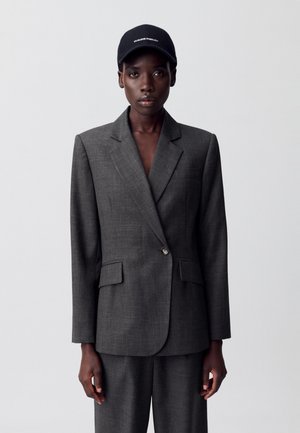 Blazer gris sur mesure avec deux poches avant, fermeture à un bouton et revers aiguisés ; porté sur un pantalon assorti et une casquette noire.